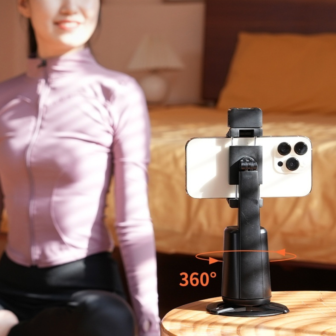 Phone holder with 360-degree rotation and face tracking on a table, blurred person in the background. 