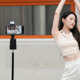 A woman dancing in front of the A200 Intelligent Face Tracking Phone Stand in black, showing AI-powered 360º rotation, gesture control, warm LED light, and built-in selfie stick for hands-free video recording.