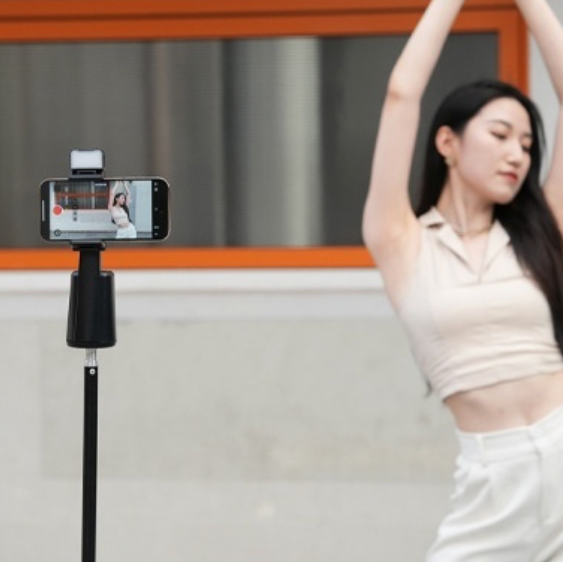A woman dancing in front of the A200 Intelligent Face Tracking Phone Stand in black, showing AI-powered 360º rotation, gesture control, warm LED light, and built-in selfie stick for hands-free video recording.