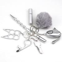 8-piece women’s self-defense keychain set in silver, including alarm, self-defense stick, window breaker, whistle, knife, knuckle tool, bottle holder, and pompom—all arranged neatly for display.