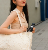 Woman holding a black and white shoulder bag against a neutral background with the A200 Intelligent Face Tracking Phone Stand in black, showing AI-powered 360º rotation, gesture control, warm LED light, and built-in selfie stick for hands-free video recording.