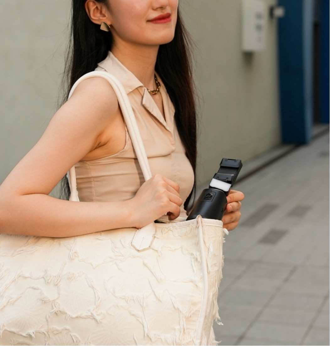 Woman holding a black and white shoulder bag against a neutral background with the A200 Intelligent Face Tracking Phone Stand in black, showing AI-powered 360º rotation, gesture control, warm LED light, and built-in selfie stick for hands-free video recording.