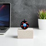 Small round hidden-camera-detector device with colored lights on a block of wood next to a laptop and plant used to detect hidden cameras