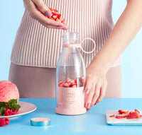 Person holding a pink bottle labeled 'Fresh Juice' with strawberries on a blue background. Portable smoothie blender in pink, compact travel blender with dual blades and USB magnetic charging, ideal for fresh smoothies on the go.