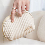 A hand holding a medium-sized beige cosmetic bag with a smooth metal zipper and a brand label reading 'WanderChics'. The bag has a shell-like design and appears to be made of PU leather.
