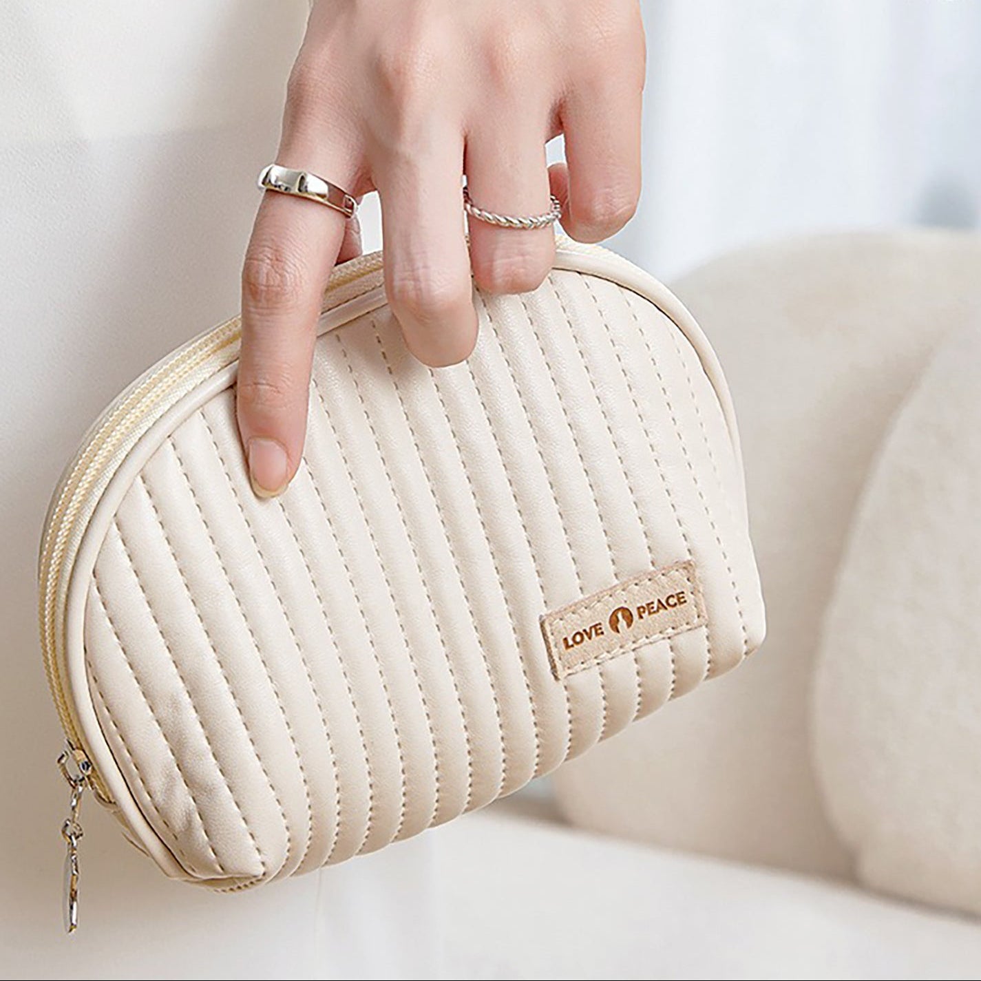 A hand holding a medium-sized beige cosmetic bag with a smooth metal zipper and a brand label reading 'WanderChics'. The bag has a shell-like design and appears to be made of PU leather.